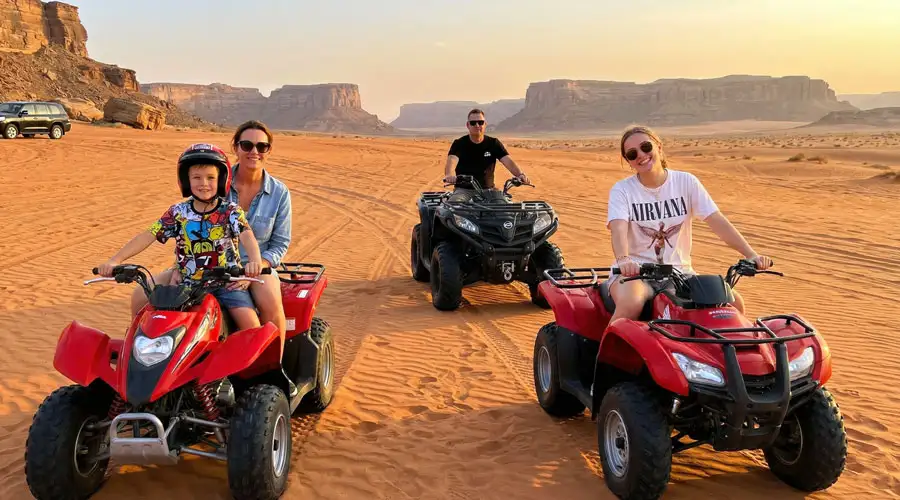 A family of four posing on red and black quad bikes in a vast desert landscape with sandstone cliffs in the background.